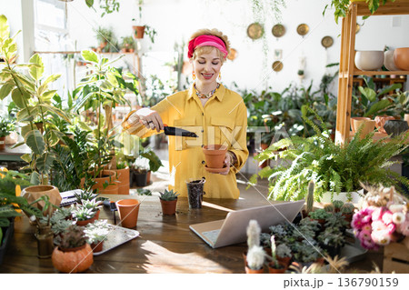 A woman in a yellow shirt and pink headscarf is planting succulents in a terracotta pot, surrounded by lush greenery A woman in a yellow shirt and pink headscarf is planting succulents in a terracotta pot, surrounded by lush greenery 136790159