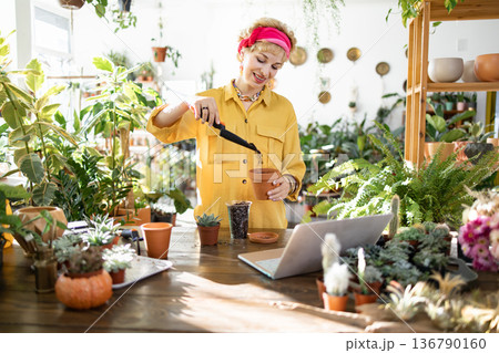 A woman smiles while repotting a plant, surrounded by lush greenery and using a laptop for guidance 136790160