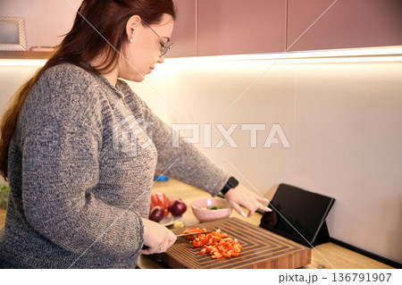 Woman controlling tablet with one hand while chopping vegetables on wooden board. 136791907