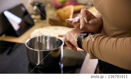 Close up of woman tapping smartwatch near pot on induction stove in modern interior. Close up of woman tapping smartwatch near pot on induction stove in modern interior. 136791911