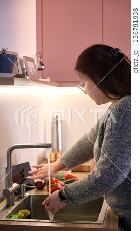 Woman washing fresh vegetables under kitchen faucet in modern home kitchen with tablet nearby. Woman washing fresh vegetables under kitchen faucet in modern home kitchen with tablet nearby. 136791938