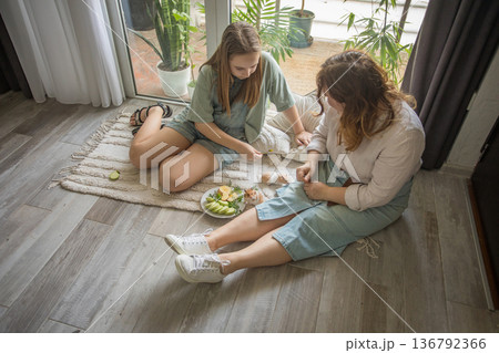 girl and women relax on a rug in a modern indoor space. They share snacks while sitting  136792366