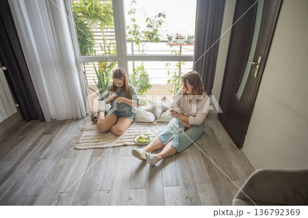 girl and women relax on a rug in a modern indoor space. They share snacks while sitting girl and women relax on a rug in a modern indoor space. They share snacks while sitting 136792369