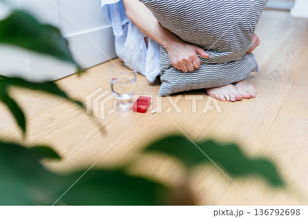 Close up of unrecognizable woman with sitting barefoot on floor, hugging pillow, glass of water and pill nearby, n, depression, mental health treatment concept at home Close up of unrecognizable woman with sitting barefoot on floor, hugging pillow, glass of water and pill nearby, n, depression, mental health treatment concept at home 136792698