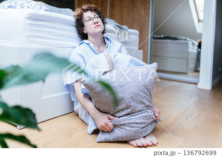 Middle aged white woman with curly hair and glasses sitting barefoot on bedroom floor, hugging pillow, looking forward with indifferent facial expression, emotional numbness, depression concept 136792699