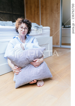 Adult woman with curly hair sitting on wooden floor in bedroom, barefoot, holding pillow close to body, direct eye contact, depression and emotional fatigue concept in private home space Adult woman with curly hair sitting on wooden floor in bedroom, barefoot, holding pillow close to body, direct eye contact, depression and emotional fatigue concept in private home space 136792700