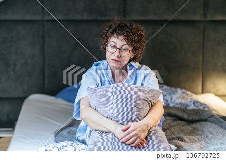 Introspective middle age woman wearing glasses sits on bed embracing pillow. Open laptop, notebook on blanket. Tired thoughtful mood, hybrid workflow pause, remote work stress, quiet bedroom interior. 136792725