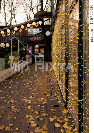 Golden leaves dance along a cozy walkway adorned with twinkling lights in autumn's gentle embrace Golden leaves dance along a cozy walkway adorned with twinkling lights in autumn's gentle embrace 136793430