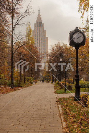 Elegant autumn stroll in a city park with a classic clock and a towering building in the backdrop Elegant autumn stroll in a city park with a classic clock and a towering building in the backdrop 136793436