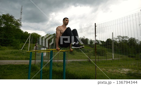 Strong guy training abdominal muscles on parallel bars at sports ground. Young shirtless sportsman exercising at nature. Sporty athlete working out at nature. Concept of active sportive lifestyle Strong guy training abdominal muscles on parallel bars at sports ground. Young shirtless sportsman exercising at nature. Sporty athlete working out at nature. Concept of active sportive lifestyle 136794344