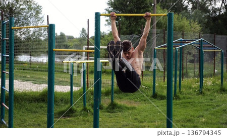 Athletic man trains abdominal muscles raising legs on horizontal bar at sports ground. Young sportsman exercising on pull-up bar outdoor. Sporty athlete working out at nature. Healthy lifestyle Athletic man trains abdominal muscles raising legs on horizontal bar at sports ground. Young sportsman exercising on pull-up bar outdoor. Sporty athlete working out at nature. Healthy lifestyle 136794345