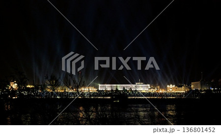golden lights illuminate waterfront scene, spectacular illumination enhances lively urban riverside spectacle, dazzling beam arrangement transforms city skyline and riverfront into vibrant celebration 136801452