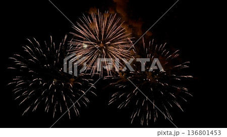 nighttime fireworks display, elegant fireworks burst over peaceful shoreline during calm evening hours, golden and bronze fireworks create balanced luminous shells above serene nighttime shoreline 136801453