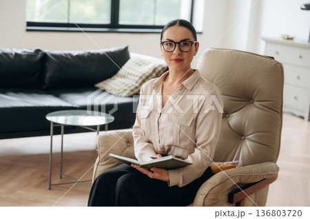 Relaxing on the chair, sitting. Woman in formal clothes is in the modern big room Relaxing on the chair, sitting. Woman in formal clothes is in the modern big room 136803720