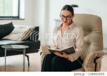 Relaxing on the chair, sitting. Woman in formal clothes is in the modern big room Relaxing on the chair, sitting. Woman in formal clothes is in the modern big room 136803959