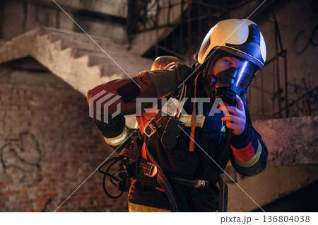 Adjusting air flow into the mask. Firefighter in professional equipment is indoors in abandoned building Adjusting air flow into the mask. Firefighter in professional equipment is indoors in abandoned building 136804038