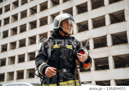 Professional firefighter is standing outdoors against big unfinished building Professional firefighter is standing outdoors against big unfinished building 136804055