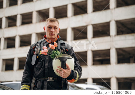 Present of flowers in hands. Professional firefighter is standing outdoors against big unfinished building Present of flowers in hands. Professional firefighter is standing outdoors against big unfinished building 136804069
