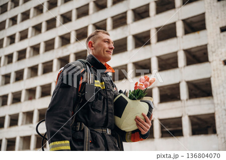 Present of flowers in hands. Professional firefighter is standing outdoors against big unfinished building Present of flowers in hands. Professional firefighter is standing outdoors against big unfinished building 136804070
