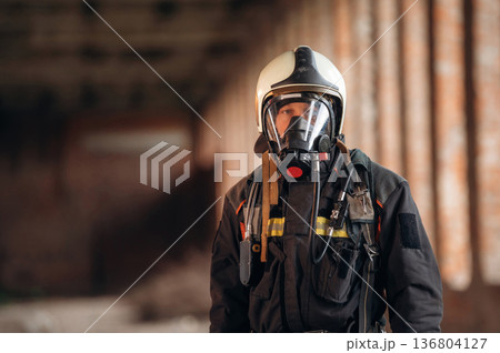 Protective air mask is on the face. Firefighter in professional equipment is indoors in abandoned building 136804127