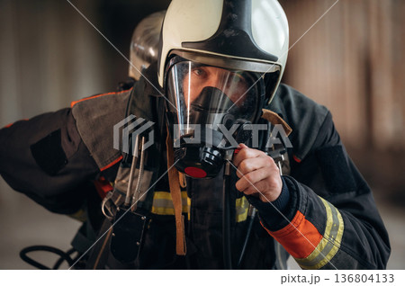 Protective air mask is on the face. Firefighter in professional equipment is indoors in abandoned building 136804133