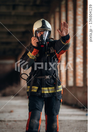 Stop hand gesture. Firefighter in professional equipment is indoors in abandoned building Stop hand gesture. Firefighter in professional equipment is indoors in abandoned building 136804139