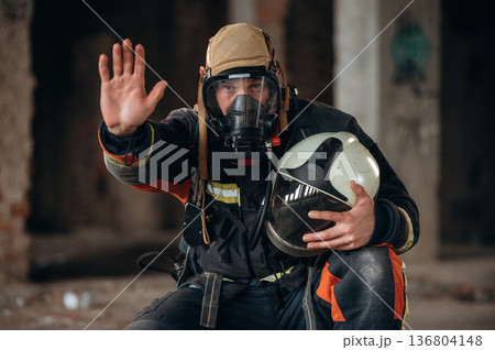 Stop hand gesture. Firefighter in professional equipment is indoors in abandoned building Stop hand gesture. Firefighter in professional equipment is indoors in abandoned building 136804148