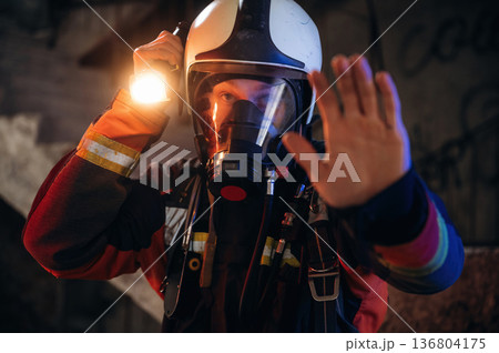 Flashlight, stop hand gesture. Firefighter in professional equipment is indoors in abandoned building 136804175