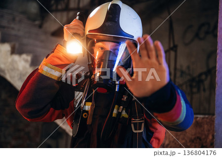 Flashlight, stop hand gesture. Firefighter in professional equipment is indoors in abandoned building Flashlight, stop hand gesture. Firefighter in professional equipment is indoors in abandoned building 136804176