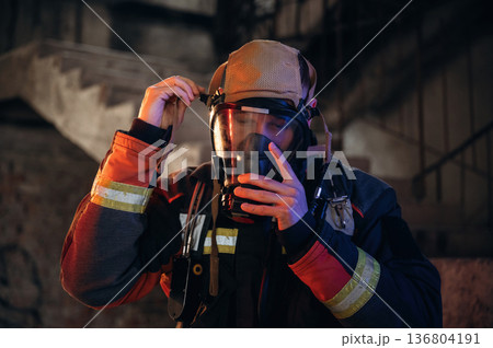 Ready, using a mask. Firefighter in professional equipment is indoors in abandoned building 136804191
