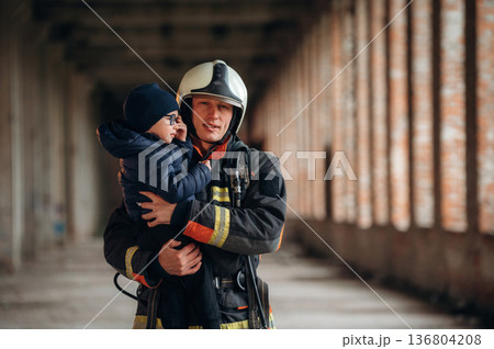Holding kid in hands. Firefighter is with baby boy indoors Holding kid in hands. Firefighter is with baby boy indoors 136804208