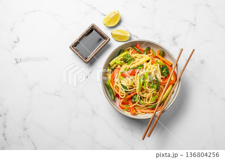 Vegetarian wok noodles with vegetables and lime in a plate on a light marble background Vegetarian wok noodles with vegetables and lime in a plate on a light marble background 136804256