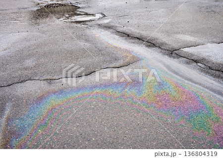 Iridescent oil slick spreading on a wet, cracked asphalt surface, reflecting light in a vibrant rainbow pattern 136804319