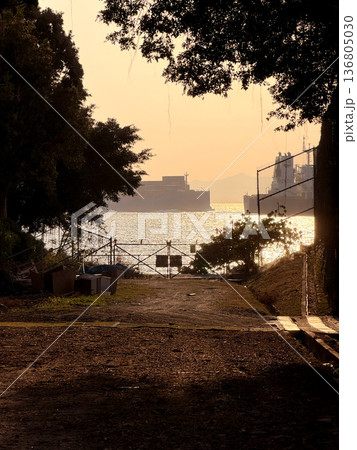Golden hour sunset over the water with cargo ships and a fenced path leading to the sea 136805030