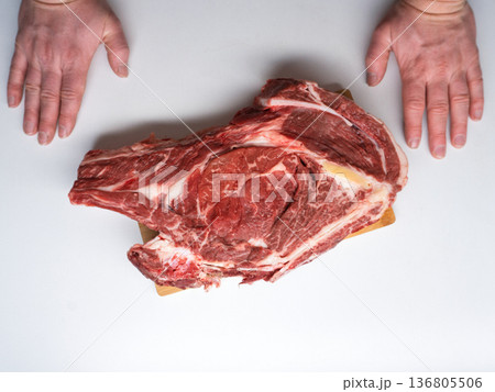 Top view of a large raw ribeye steak on a wooden board with male hands. Fresh beef meat flat lay on white background. Copy space 136805506