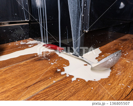 Spilled milk puddle on wooden floor with metal ladle. White liquid splashing on black kitchen cabinets. Messy accident concept 136805507