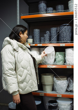 Vertical photo of a woman shopping for flower pots in a retail store. Female customer in a winter coat choosing a white planter from a shelf in a garden center 136805512