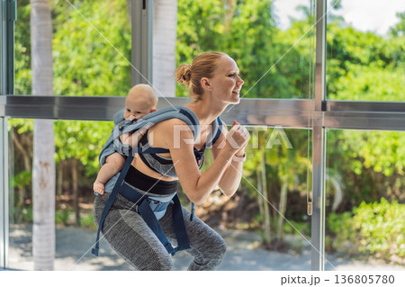 Mother doing fitness exercises while holding her baby at home, combining workout and childcare. Real lifestyle moment showing balance, strength and love. Postpartum fitness, healthy motherhood and 136805780
