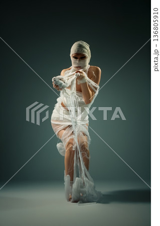 A woman wrapped in white bandages like a mummy, posing dramatically in a studio. 136805910