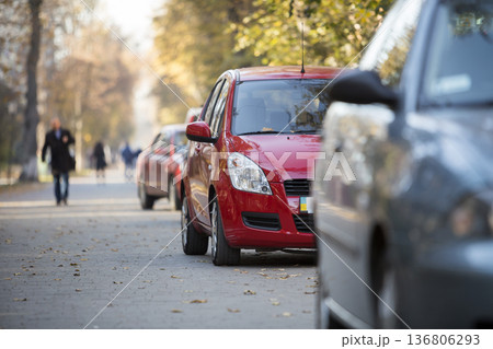 Close-up front view detail of bright red shiny car parked on pavement on background of blurred silhouette of walking people on sunny summer day. Modern city lifestyle, parking problems concept. Close-up front view detail of bright red shiny car parked on pavement on background of blurred silhouette of walking people on sunny summer day. Modern city lifestyle, parking problems concept. 136806293