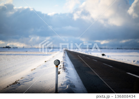 雪原をまっすぐ伸びる冬の田んぼ道：ポツンと立つ視線誘導標と青空 136808434