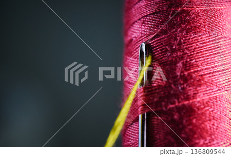 Macro shot of a metal needle eye with yellow thread against a red textile thread spool 136809544