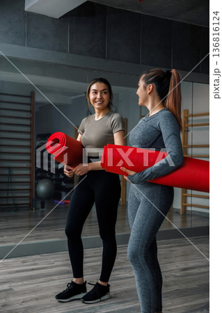 Two women hold red yoga mats in a gym while preparing for training and discussing their fitness plans 136816124