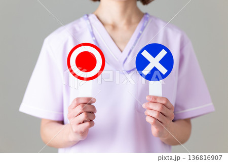 丸バツのプレートを持って悩む医療用スクラブ姿のミドル女性看護師 136816907