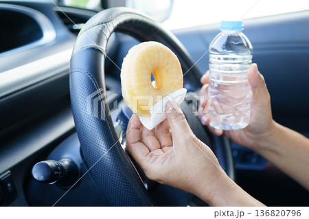 Bangkok, Thailand March 16, 2025 Asian woman driver hold cold water and sandwich bread for eat and drink in car, dangerous and risk an accident. 136820796
