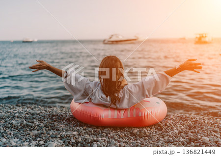 Woman beach ocean. Relaxing woman sitting on pebble beach, inflatable ring, embracing vast ocean view at sunrise. Woman beach ocean. Relaxing woman sitting on pebble beach, inflatable ring, embracing vast ocean view at sunrise. 136821449
