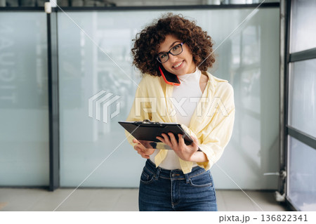 Writing the information while talking by the phone. Beautiful woman is standing indoors against blurred wall 136822341
