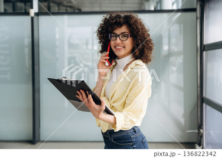 Writing the information while talking by the phone. Beautiful woman is standing indoors against blurred wall 136822342