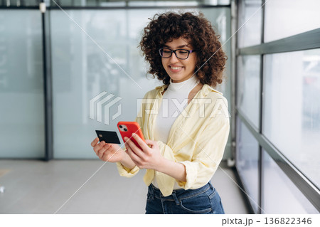 Credit card and phone in hands. Beautiful woman is standing indoors against blurred wall 136822346