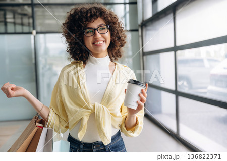 Cup of drink and shopping bags are in hands. Beautiful woman is standing indoors against blurred wall Cup of drink and shopping bags are in hands. Beautiful woman is standing indoors against blurred wall 136822371